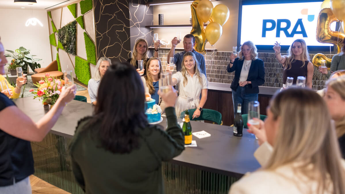 A large group of PRA employees raises glasses in a toast beneath gold balloons and a screen displaying the PRA logo, celebrating the company's anniversary.