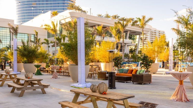 Outdoor lounge area with picnic tables, string lights, and palm trees at dusk.