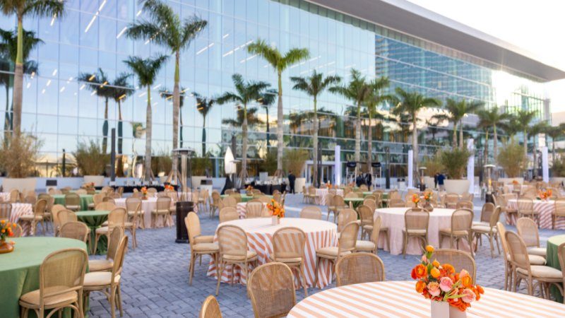 Round tables with orange-striped linens and floral centerpieces in front of a glass building lined with palm trees.