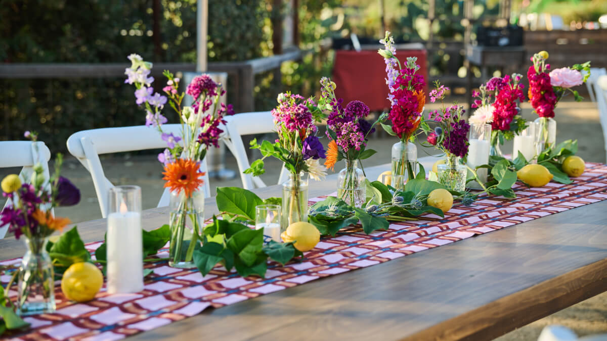 Close-up of a long wooden table decorated with colorful flowers in glass vases and lemons arranged along a patterned runner.