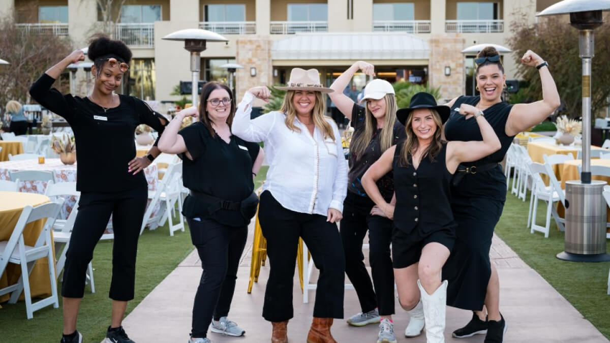 Six women pose playfully flexing their arms outdoors at an evening event with round tables, gold linens, and string lighting in the background.
