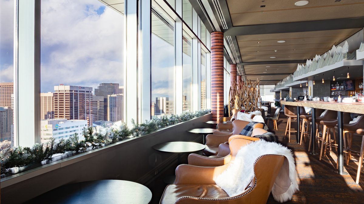 A high-rise lounge with floor-to-ceiling windows overlooking a city skyline, leather armchairs draped with faux fur throws, evergreen garland along the windowsill, and a long bar.