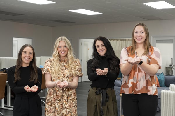 Four women in an office setting smiling with open hands extended, symbolizing sharing andGive to gain