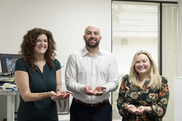Three coworkers standing side by side with open hands, smiling and reflecting the concept ofGive to gain