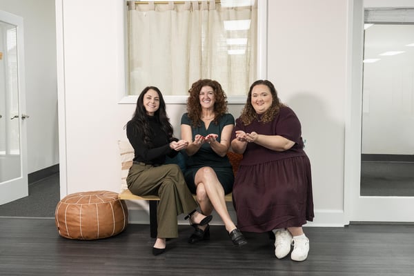 Three women seated on a bench, smiling and holding their hands open as a gesture of giving and receiving.