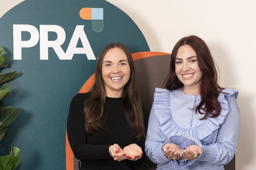 Two smiling women standing in front of a PRA backdrop, symbolizing the idea of Give to gain.