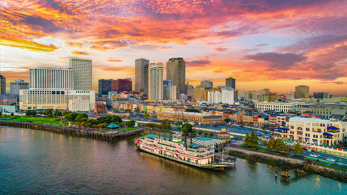 Aerial view of downtown New Orleans skyline and the Mississippi River riverfront at sunset, with a paddlewheel riverboat docked near the French Quarter.