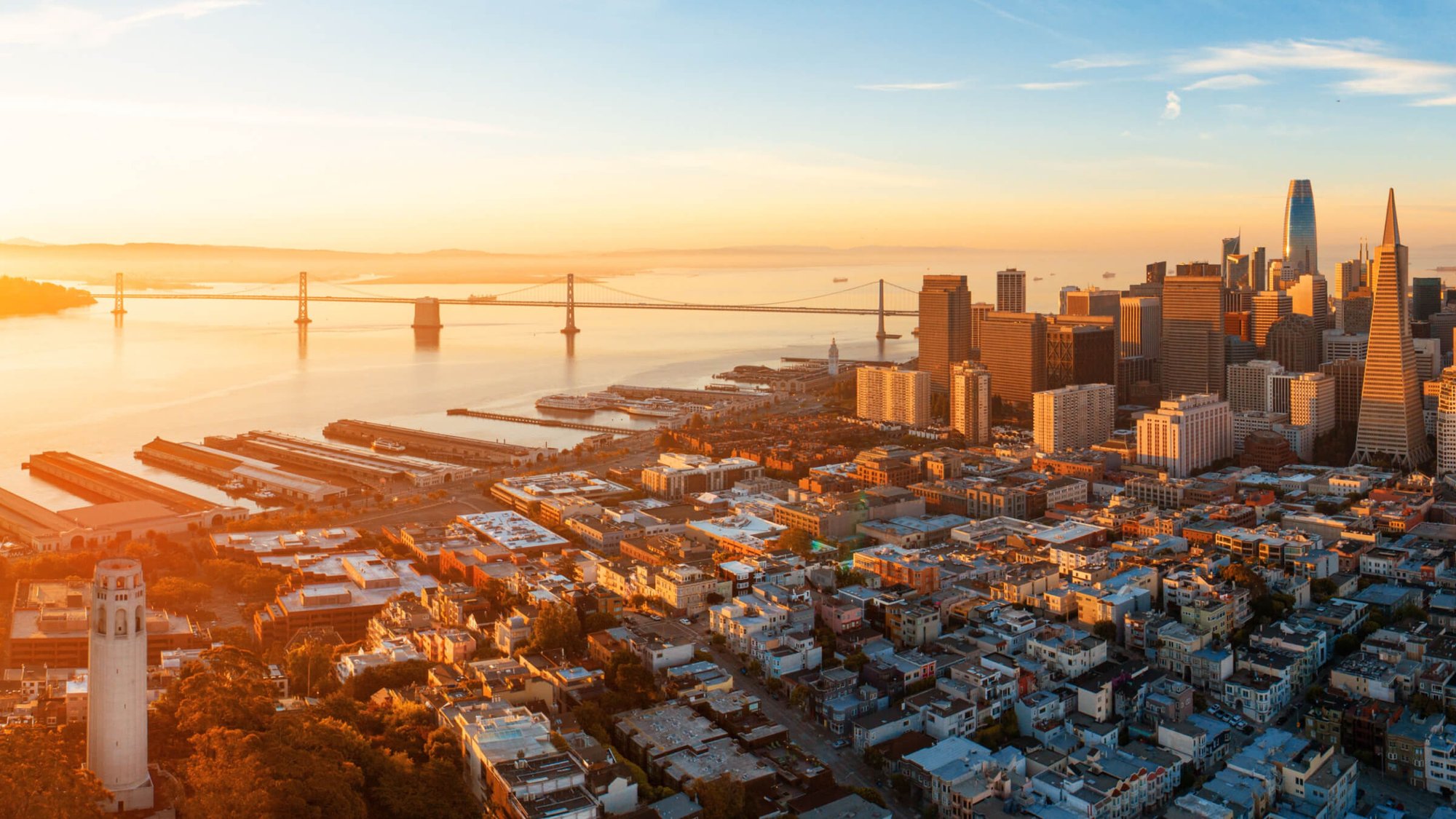 Aerial view of San Francisco's skyline and waterfront at sunset, with the Bay Bridge, Coit Tower, and downtown high-rises visible.