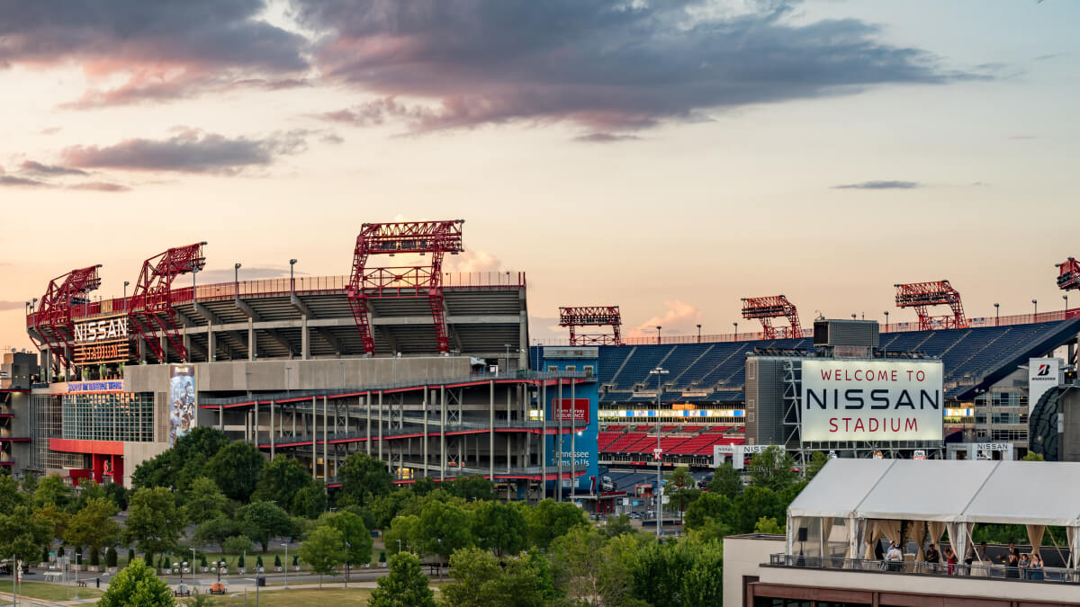 Sunset view of Nissan Stadium in Nashville, home of the Tennessee Titans, with red gantry structures, a \"Welcome to Nissan Stadium\" sign, and a rooftop event space in the foreground.