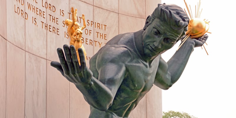 Statue of the Spirit of Detroit holding a golden sphere, with engraved text on a stone wall behind.