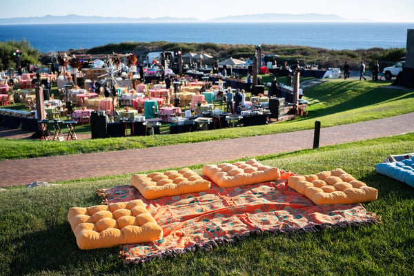 Lounge seating with cushions arranged on grass overlooking a large outdoor event by the ocean.