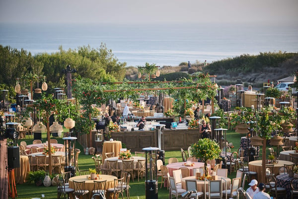 Wide view of an outdoor event space with dining tables and greenery near the coastline.