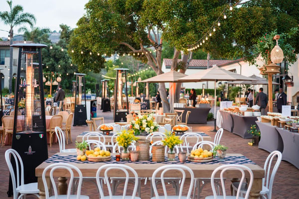 Long outdoor dining table with white chairs, floral décor, and patio heaters under string lights.