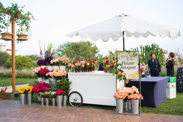 Outdoor flower stand with colorful blooms and a white umbrella in a garden setting.