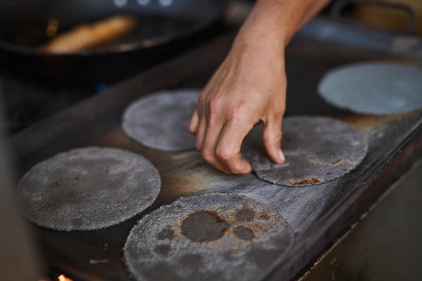 Close-up of a cook’s hand flipping corn tortillas on a hot griddle during food preparation.