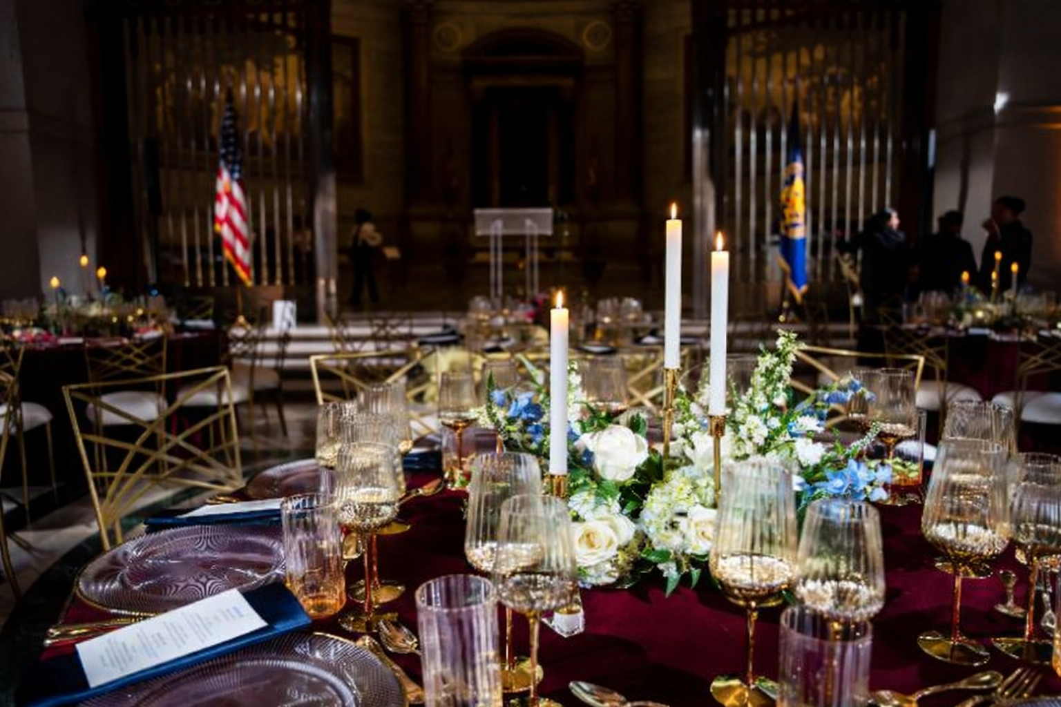 Formal banquet table with candles, glassware, and floral centerpiece in a dimly lit venue with flags in the background.