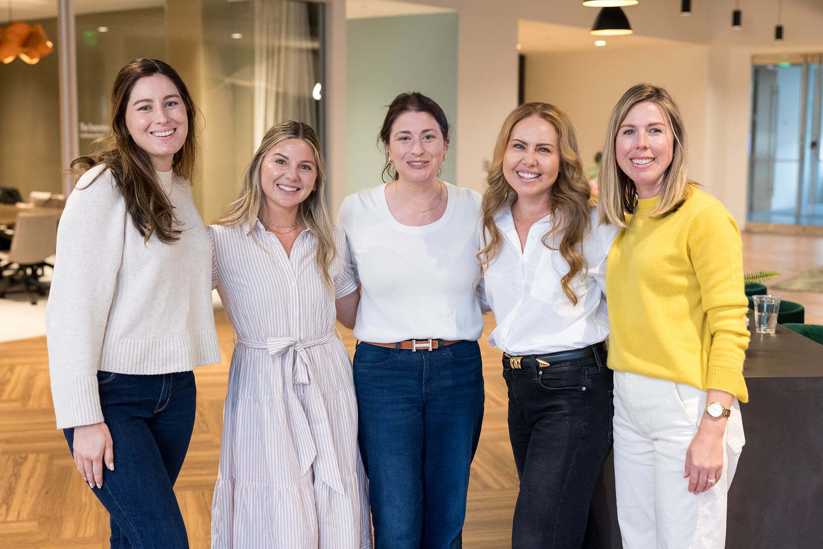 Five women stand arm-in-arm smiling for a group photo in a modern office lounge.