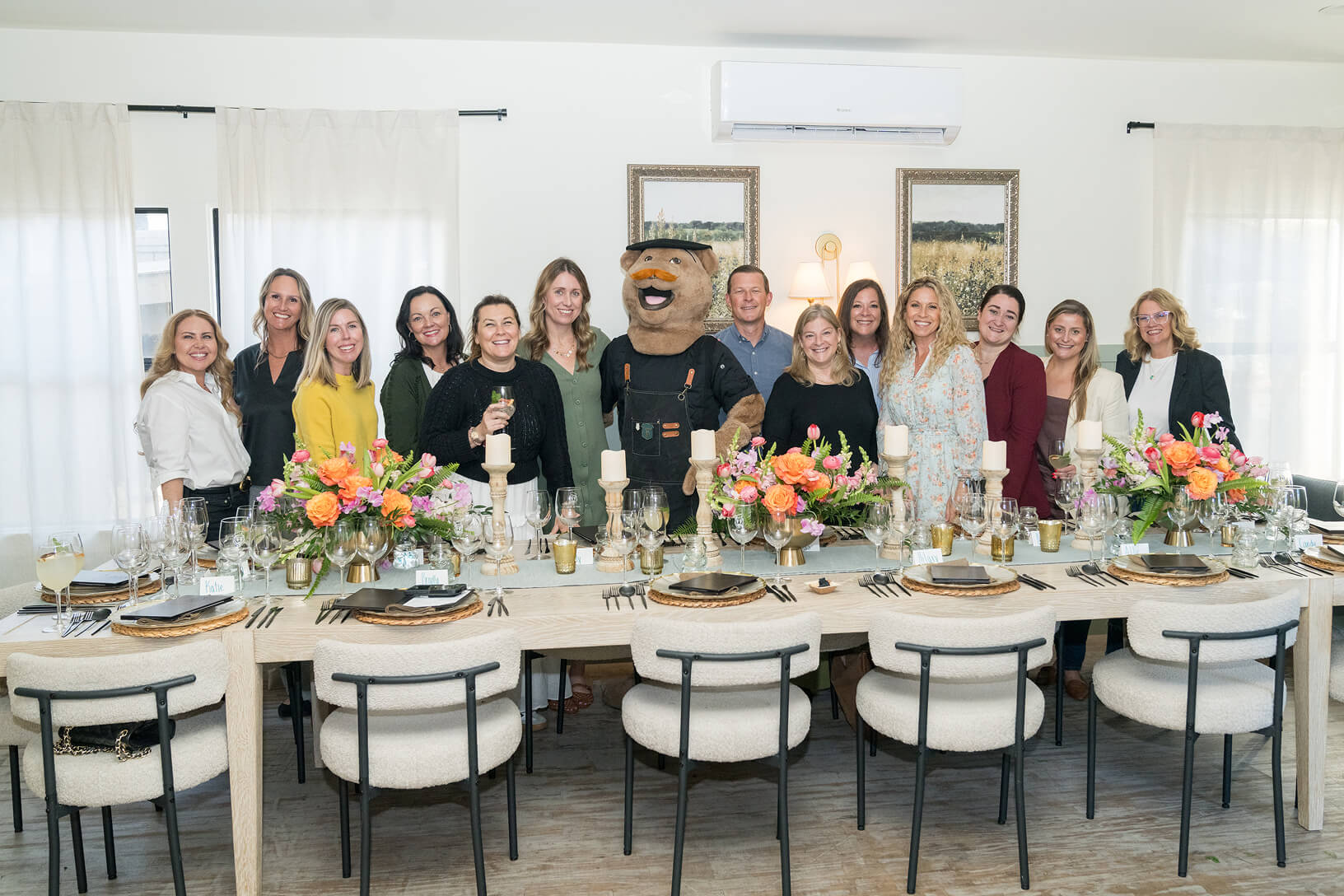 A group of PRA colleagues stand behind a long dinner table decorated with floral centerpieces and candles, joined by a costumed bear mascot, celebrating together at the Aged to Perfection dinner.