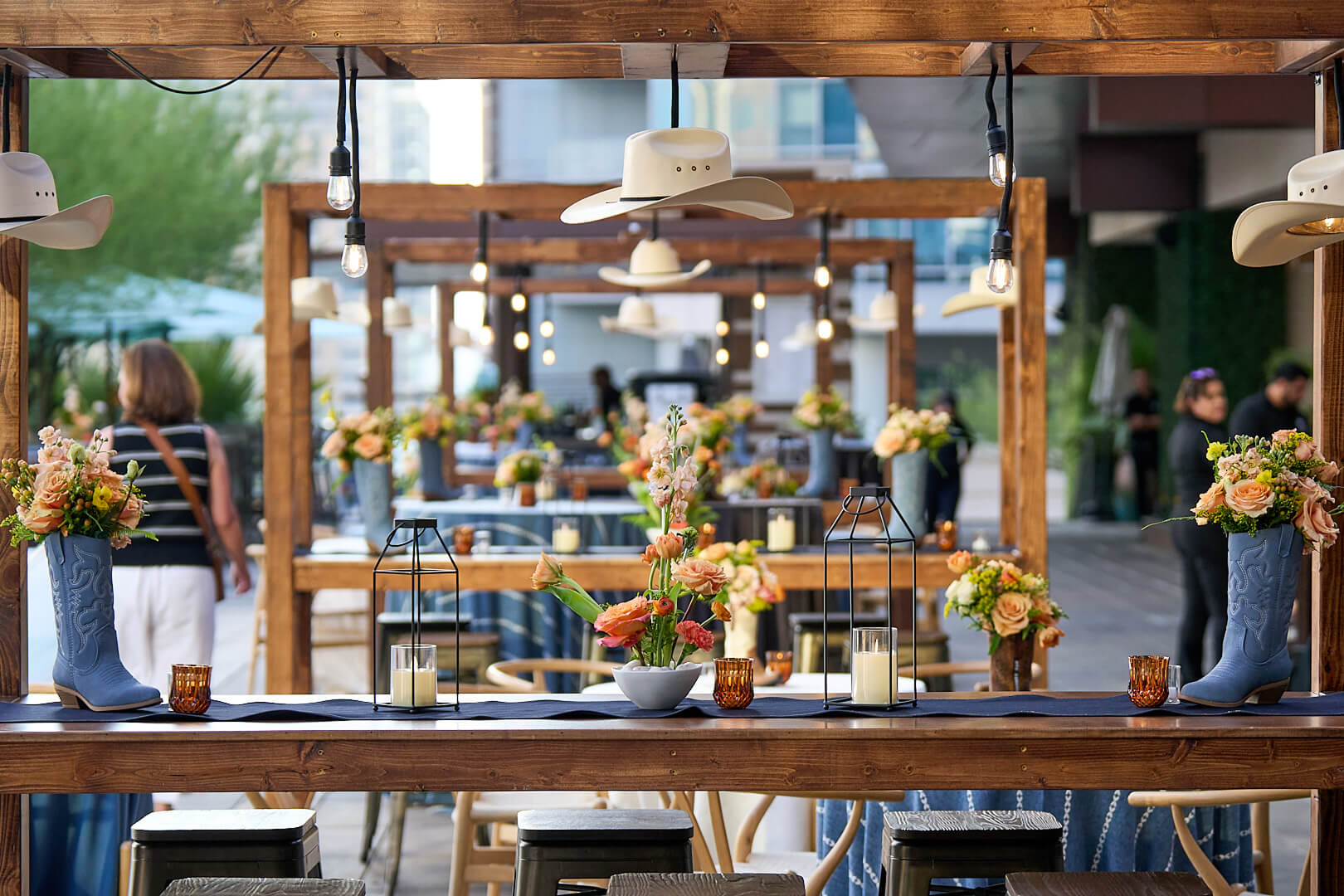 Rustic dining table decorated with flowers, lanterns, and cowboy hats in an outdoor event space.