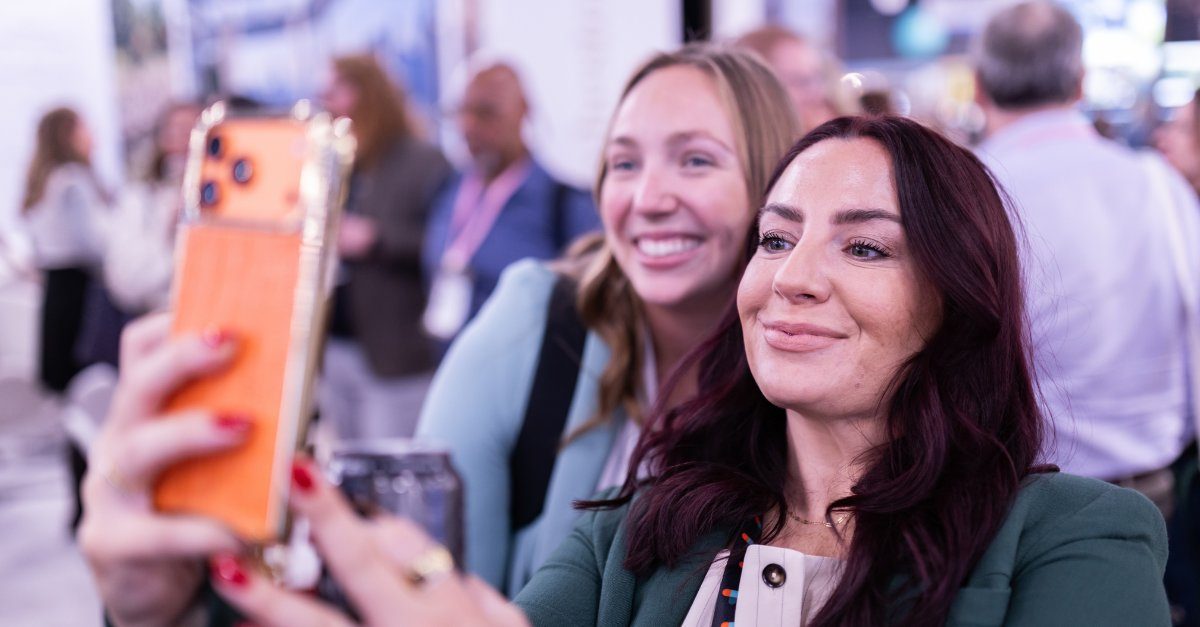 Two attendees smiling while taking a selfie at a busy conference expo.