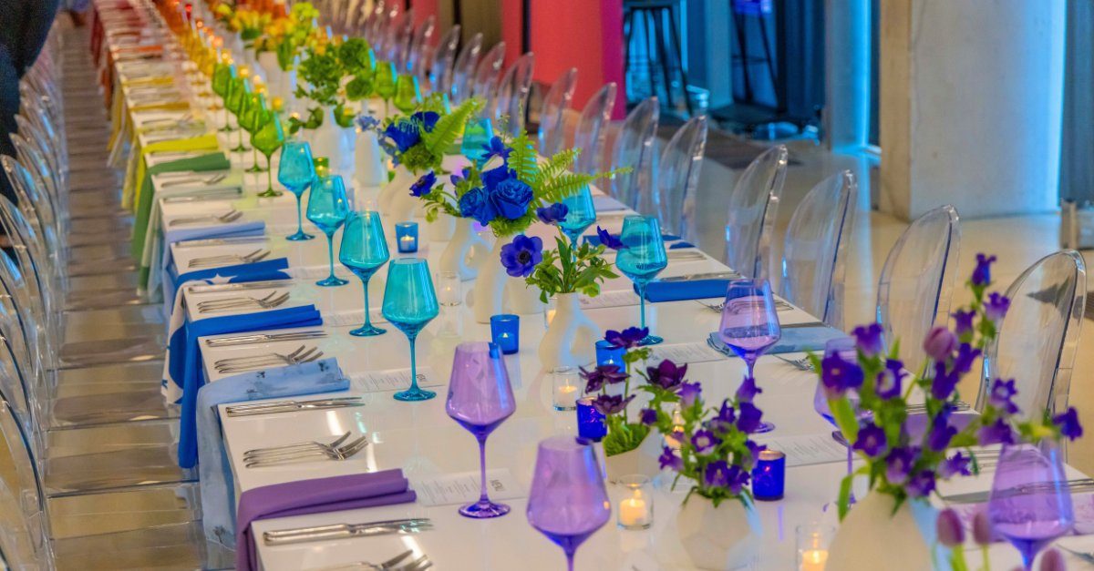 Long banquet table set with a rainbow gradient of colored glassware, linens, and flowers.
