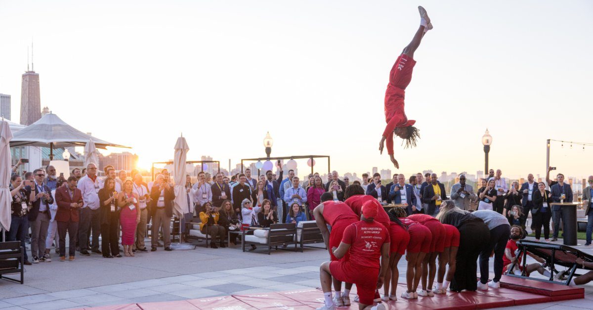A performer flips through the air above a human pyramid at an outdoor rooftop event as guests watch.