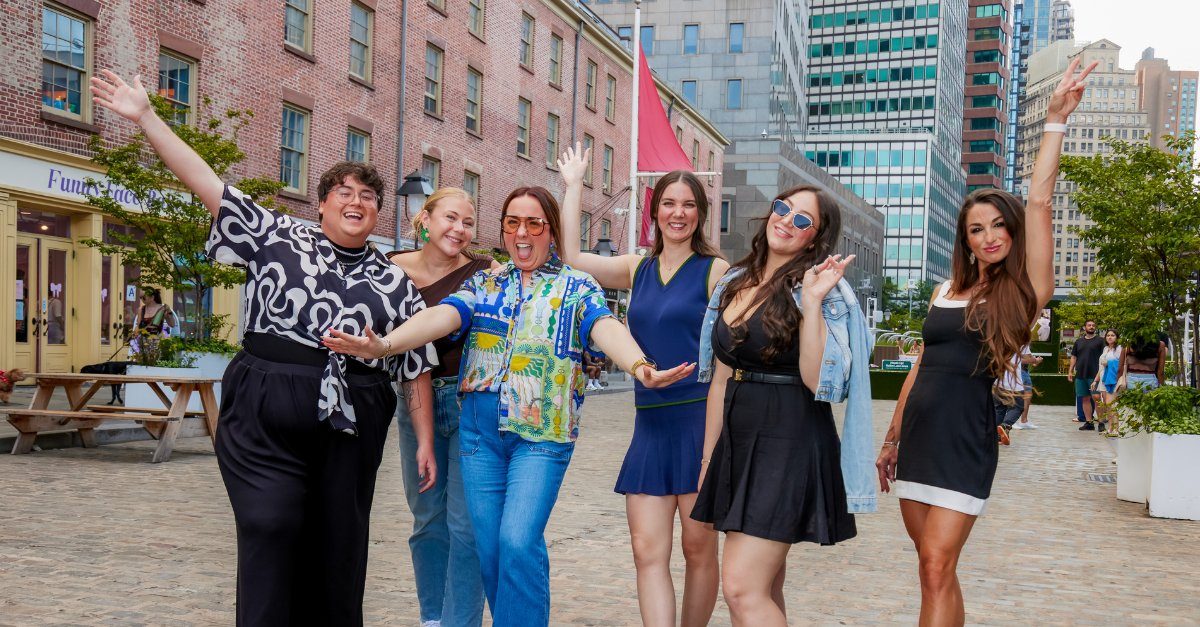 A group of six friends laughing and posing with raised arms on a city street.