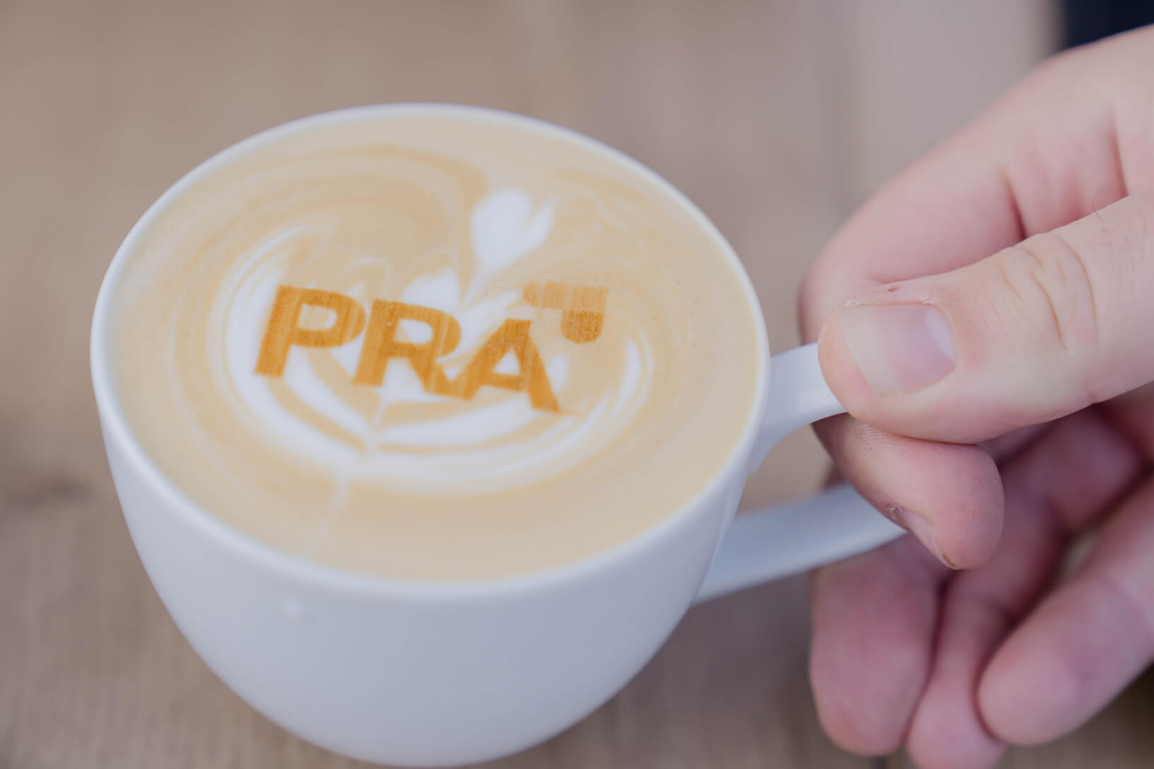 A hand holds a cup of latte decorated with the PRA logo in the foam.