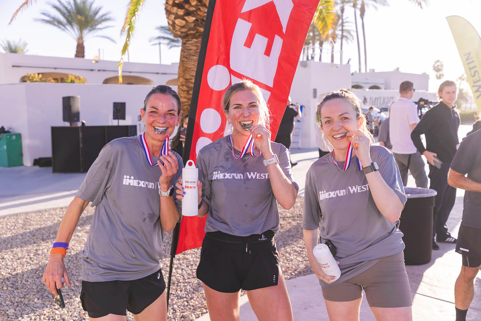 Three IMEXrun participants smile and bite their finisher medals beside an IMEX flag in Las Vegas.