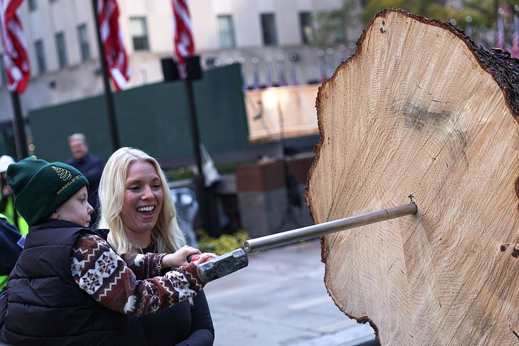 A smiling woman and a young child in a green beanie hammer a spike into the cut trunk of a giant evergreen at Rockefeller Plaza.