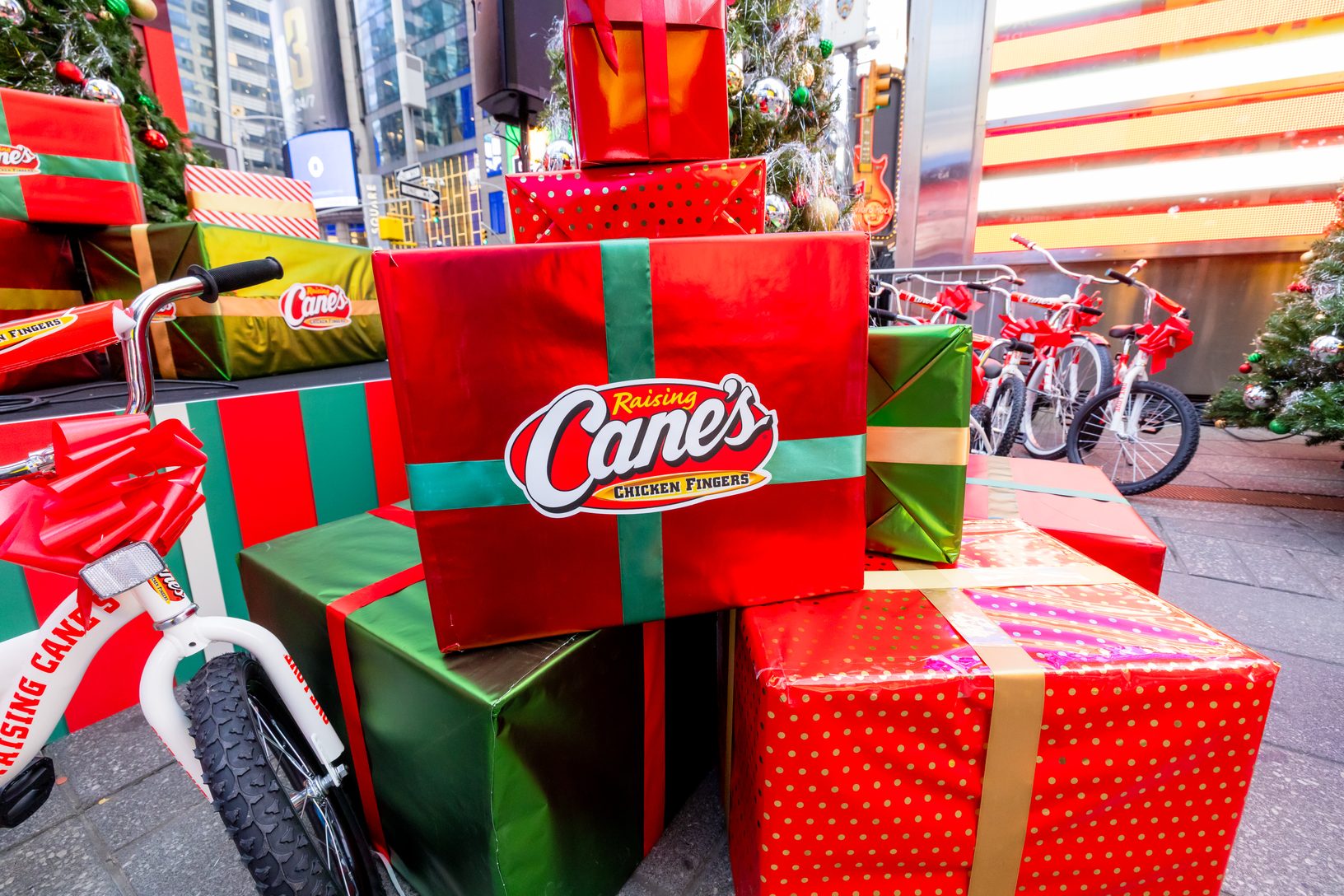 Stacked red and green gift boxes with Raising Cane's branding in Times Square, beside a white kids' bike and rows of bicycles tied with red bows.