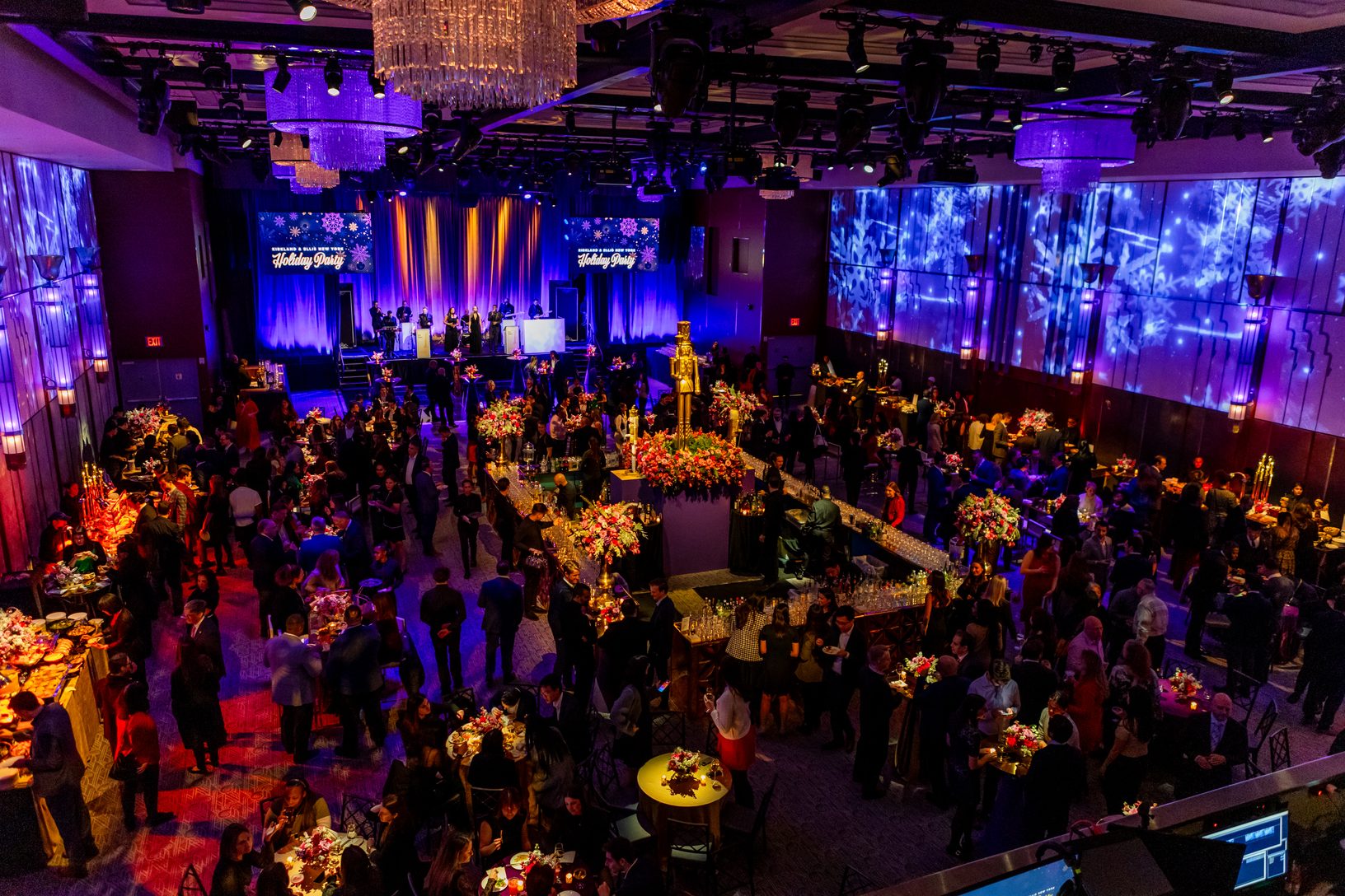 Overhead view of a packed holiday party in a ballroom with purple lighting, snowflake projections, crystal chandeliers, a live band and a gold nutcracker centerpiece over a central bar.