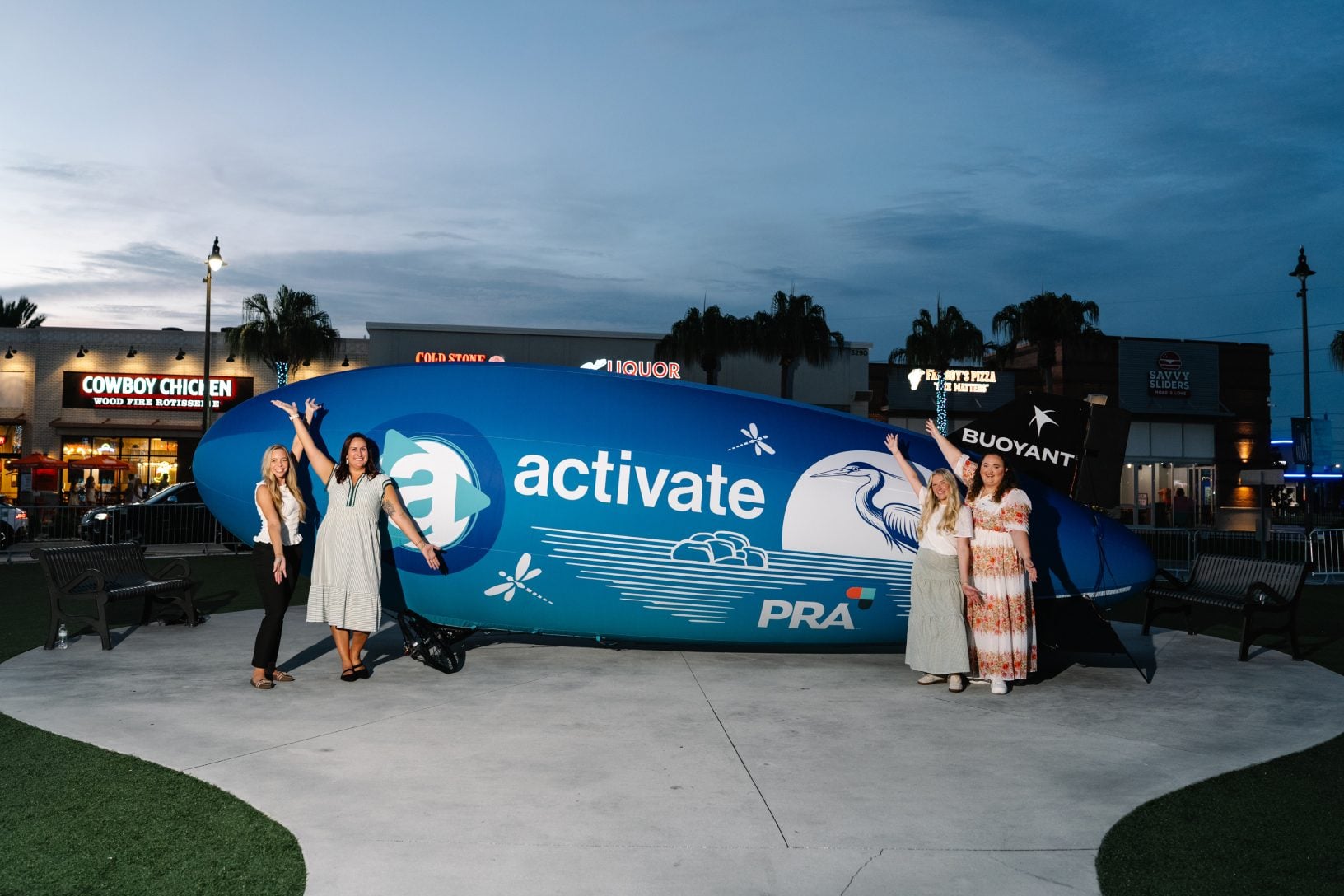 Four event partners pose beside the large custom Maritz Activate and PRA blimp at Sunset Walk after dark.