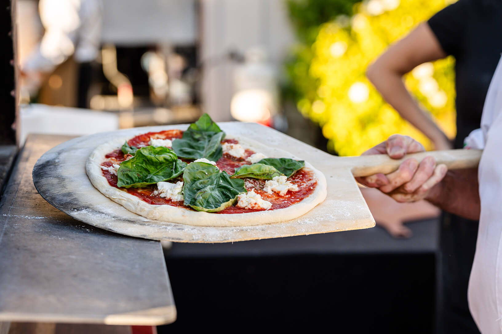 A chef slides a fresh basil and mozzarella pizza off a wooden peel at an outdoor event food station.