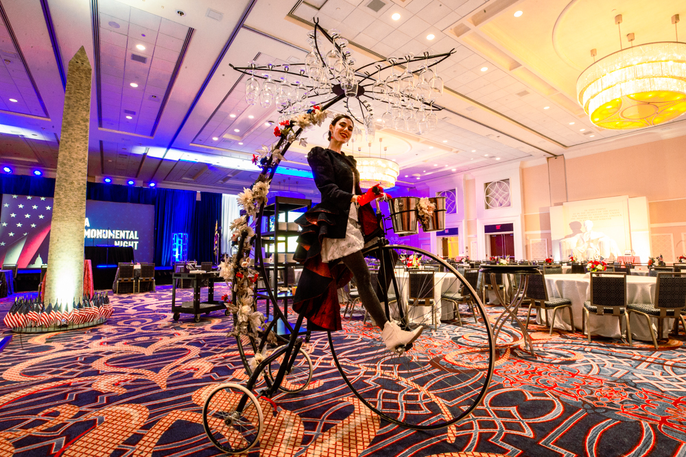 Performer riding a decorative high-wheel bicycle inside a ballroom with stage lighting.